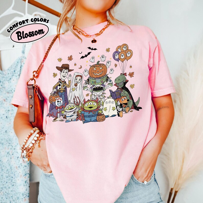 May include: Pale pink t-shirt with a Halloween cartoon graphic. The design features characters in costumes, including a pumpkin, ghosts, and bats. The text "Comfort Colors Blossom" is printed on the upper left.