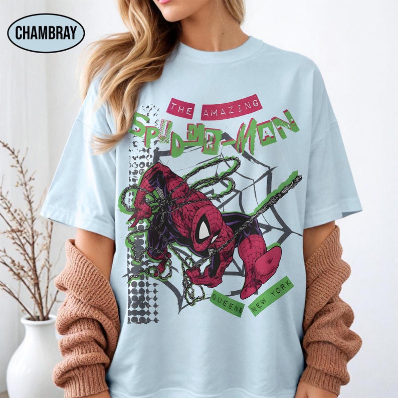 May include: Light blue t-shirt featuring a graphic of Spider-Man in a web, with text reading "THE AMAZING SPIDER-MAN" in green and red. The design includes the words "QUEENS NEW YORK". The shirt has a relaxed fit.
