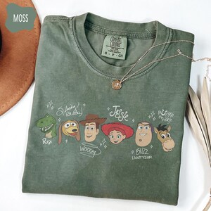 May include: A moss green crew neck t-shirt featuring a graphic print of six animated characters with their names written in a script font: Rex, Slinky Dog, Woody, Jessie, Buzz Lightyear, and Bullseye the Horse. The shirt has a Comfort Colors label.