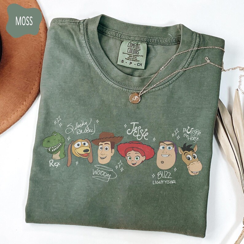 May include: A moss green crew neck t-shirt featuring a graphic print of six animated characters with their names written in a script font: Rex, Slinky Dog, Woody, Jessie, Buzz Lightyear, and Bullseye the Horse. The shirt has a Comfort Colors label.