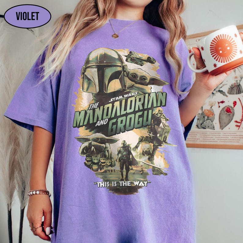 May include: Violet-colored t-shirt featuring a graphic from the Star Wars series, "The Mandalorian and Grogu." The design includes images of the Mandalorian, Grogu, and other characters, with the phrase "THIS IS THE WAY" at the bottom.