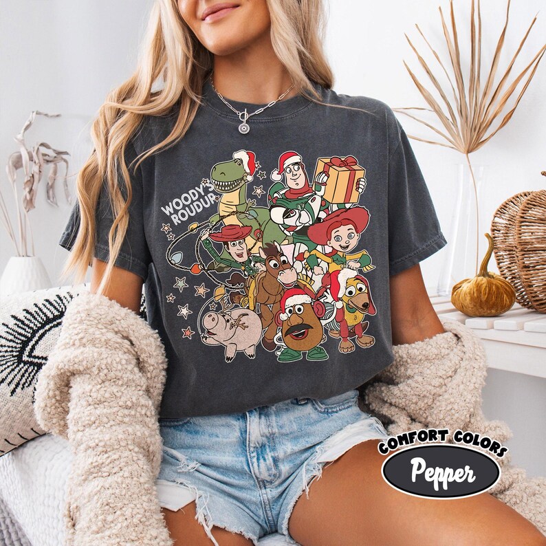 Op de afbeelding: Donkergrijs t-shirt met een afbeelding van Toy Story-personages met kerstmutsen. De tekst "Woody's Roundup" en "Comfort Colors Pepper" staat op het shirt. Het ontwerp bevat Woody, Buzz Lightyear en andere personages.