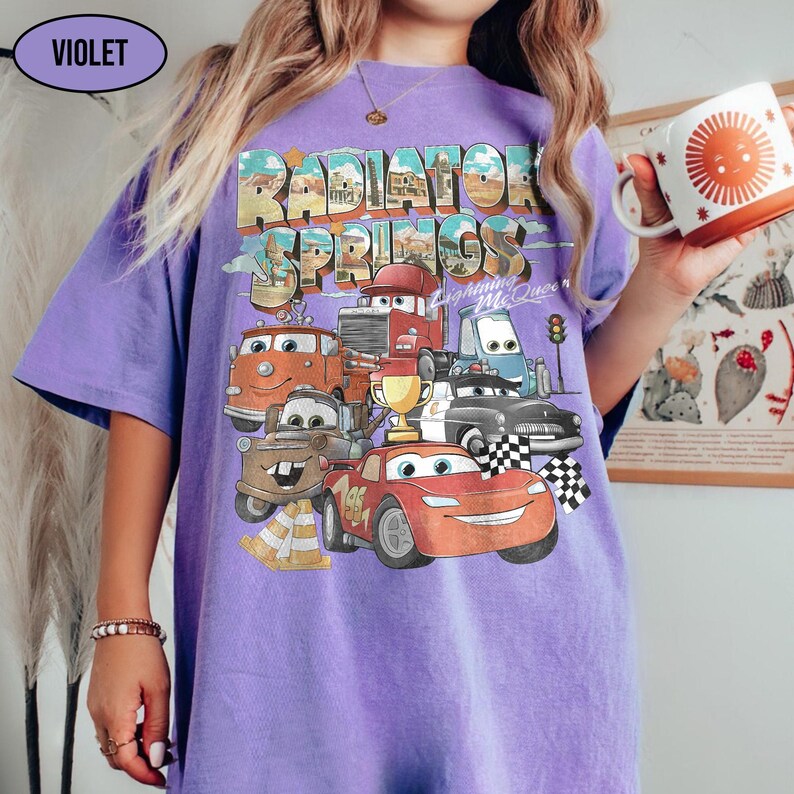 May include: A violet short-sleeve t-shirt featuring a graphic print of Radiator Springs with Lightning McQueen and other characters from the movie Cars. The design includes a gold trophy, traffic cones, and checkered flags with text.