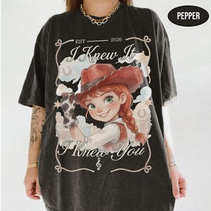 May include: A charcoal grey t-shirt featuring a graphic of a cartoon cowgirl with red hair in braids and a red cowboy hat. The text reads "EST 2026", "I Knew It", and "I Knew You" with a rope border. A small black oval in the corner says "PEPPER".