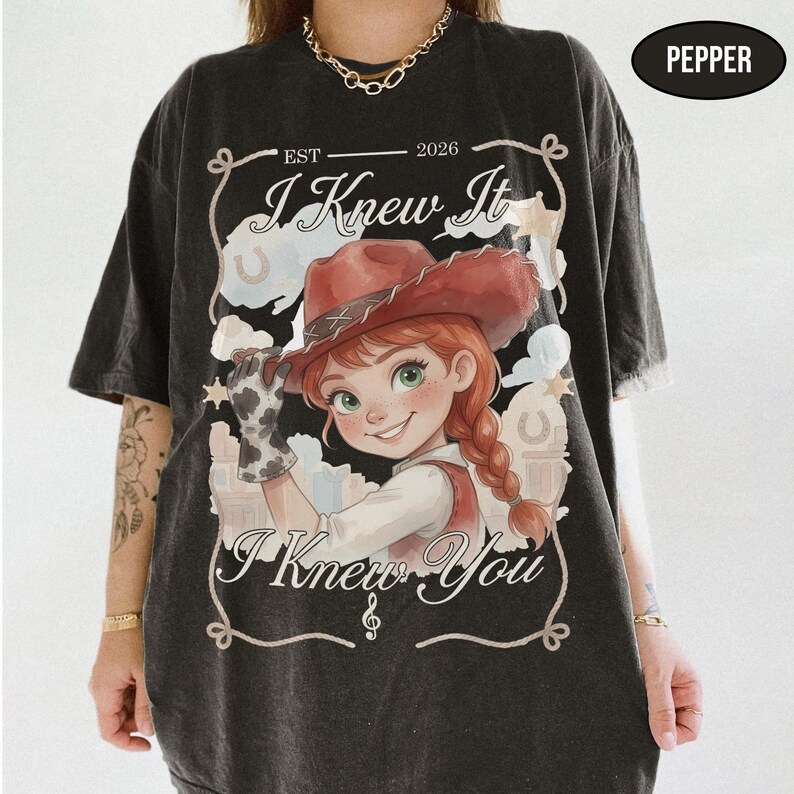 May include: A charcoal grey t-shirt featuring a graphic of a cartoon cowgirl with red hair in braids and a red cowboy hat. The text reads "EST 2026", "I Knew It", and "I Knew You" with a rope border. A small black oval in the corner says "PEPPER".