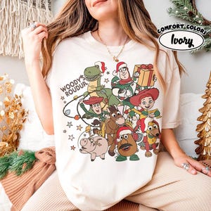 Op de afbeelding: Ivoor kleurig T-shirt met een feestelijke afbeelding van Toy Story-personages met kerstmutsen. Het ontwerp bevat Woody, Buzz Lightyear, Rex en andere personages, met de tekst "Woody's Roundup". Het shirt is gemarkeerd met "Comfort Colors".
