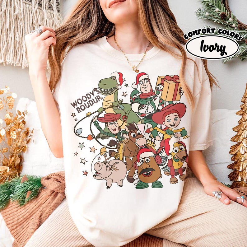 Op de afbeelding: Ivoor kleurig T-shirt met een feestelijke afbeelding van Toy Story-personages met kerstmutsen. Het ontwerp bevat Woody, Buzz Lightyear, Rex en andere personages, met de tekst "Woody's Roundup". Het shirt is gemarkeerd met "Comfort Colors".