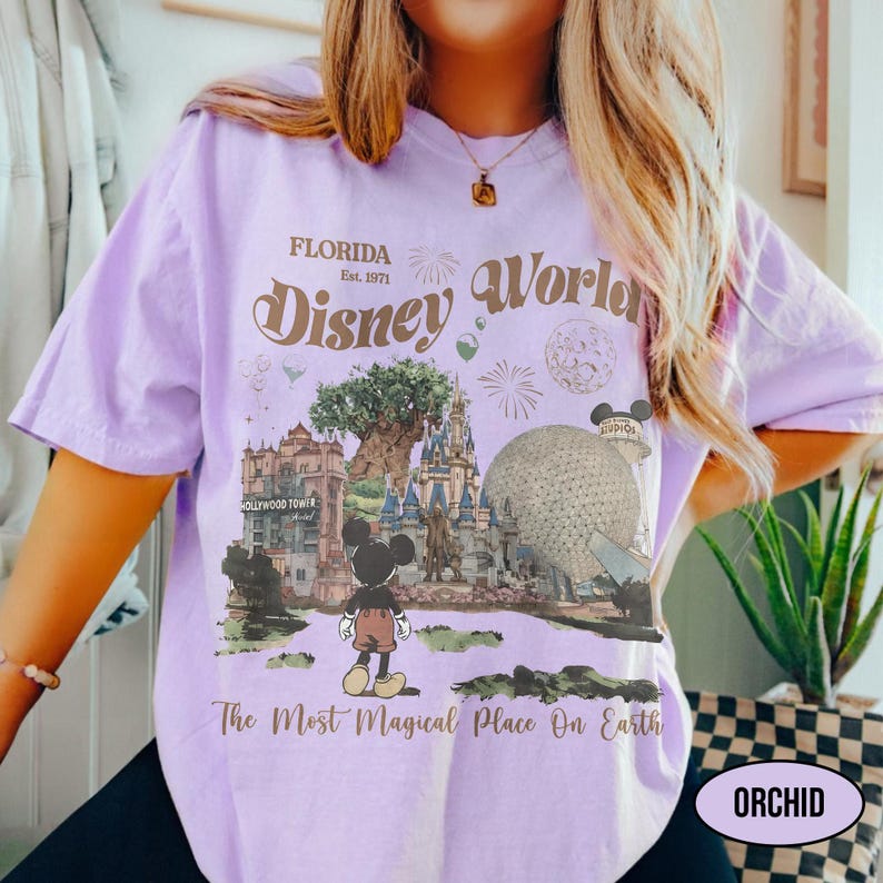Puede incluir: Camiseta de color lila claro con un dise&ntilde;o de Disney World en Florida. El dise&ntilde;o incluye el texto "FLORIDA, Est. 1971", "Disney World" y "The Most Magical Place On Earth". La camiseta tambi&eacute;n tiene una peque&ntilde;a etiqueta "ORCHID".