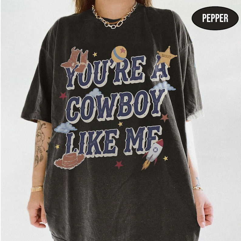 Puede incluir: Una camiseta de manga corta gris carb&oacute;n con el texto YOU'RE A COWBOY LIKE ME en letras azules. El gr&aacute;fico incluye ilustraciones de botas de vaquero marrones, una pelota, una estrella de sheriff, nubes, un sombrero marr&oacute;n, un cohete y estrellas rojas.