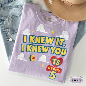 Puede incluir: Una camiseta de manga corta color morado claro con el texto I KNEW IT, I KNEW YOU en amarillo con bordes azules. El dise&ntilde;o incluye nubes blancas, estrellas amarillas, un coraz&oacute;n rojo con TS STORY 5 en amarillo y una pelota amarilla con estrella roja.