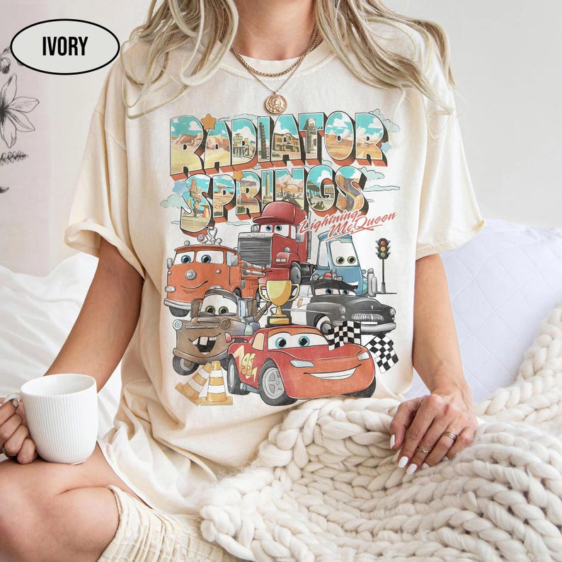 May include: An ivory t-shirt featuring a graphic print of Radiator Springs with various cartoon cars, including a red race car, a brown tow truck, and a gold trophy. The text reads Radiator Springs and Lightning McQueen in a retro font.