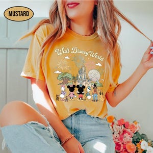 May include: Mustard-coloured t-shirt with a graphic of Disney characters in front of Cinderella's castle and the text "Walt Disney World". The shirt has a retro design with fireworks and clouds.