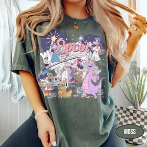 May include: Moss green t-shirt featuring a colorful graphic of Disney characters painting the word "Epcot". The design includes Mickey Mouse, Minnie Mouse, Donald Duck, and a purple dragon. The shirt has a vintage, slightly faded look.