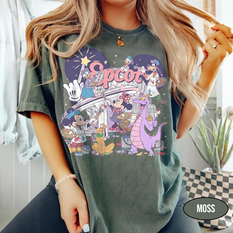 May include: Moss green t-shirt featuring a colorful graphic of Disney characters painting the word "Epcot". The design includes Mickey Mouse, Minnie Mouse, Donald Duck, and a purple dragon. The shirt has a vintage, slightly faded look.