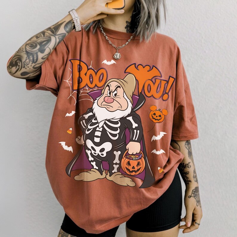 May include: A rust-colored t-shirt featuring a graphic of Grumpy from Snow White dressed in a skeleton costume, holding a jack-o'-lantern bucket. The design includes the text "Boo You!", spiderwebs, bats, and candy corn on a solid background.