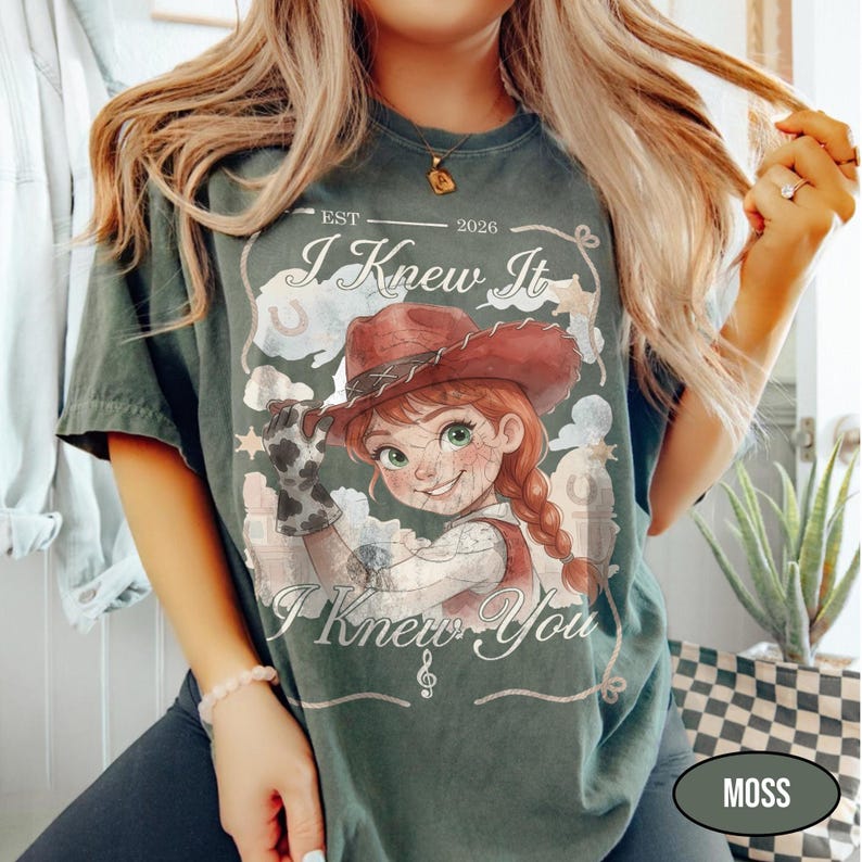 May include: A dark green t-shirt featuring a graphic of a cartoon cowgirl in a red hat and cow-print gloves. The text reads EST 2026, I Knew It, and I Knew You, accompanied by a musical note symbol, stars, and horseshoe illustrations.