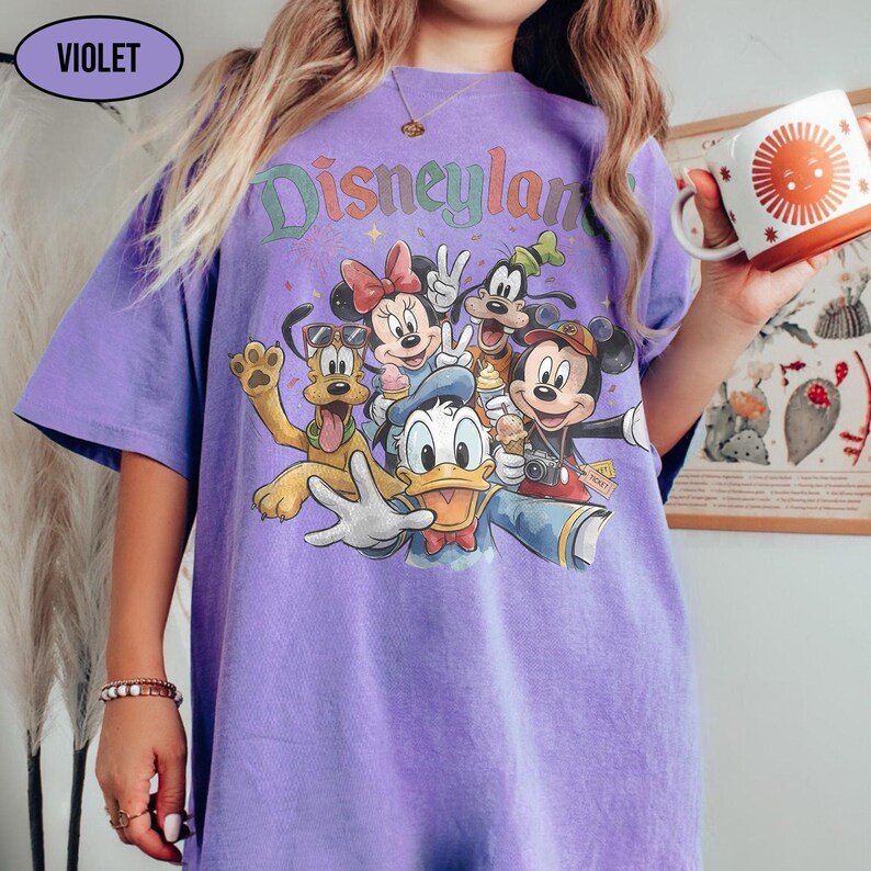 May include: A violet-colored t-shirt featuring a graphic of Disney characters, including Mickey Mouse, Minnie Mouse, Donald Duck, Goofy, and Pluto. The word "Disneyland" is printed across the top in a colorful, retro font. The shirt is a short-sleeved, oversized style.