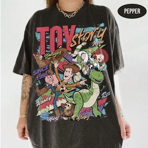 Puede incluir: Una camiseta gris oscuro con un gr&aacute;fico colorido de Toy Story, con personajes como Woody y Buzz Lightyear. Se muestra el texto "TOY STORY" y "SINCE 1995". La camiseta tiene un aspecto vintage.