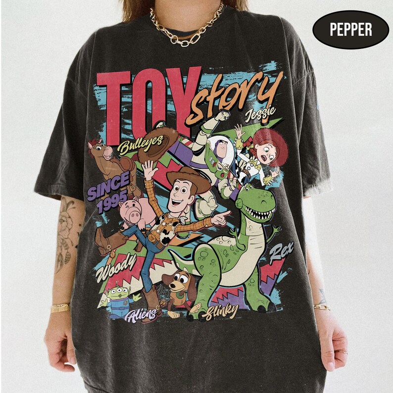 Puede incluir: Una camiseta gris oscuro con un gr&aacute;fico colorido de Toy Story, con personajes como Woody y Buzz Lightyear. Se muestra el texto "TOY STORY" y "SINCE 1995". La camiseta tiene un aspecto vintage.