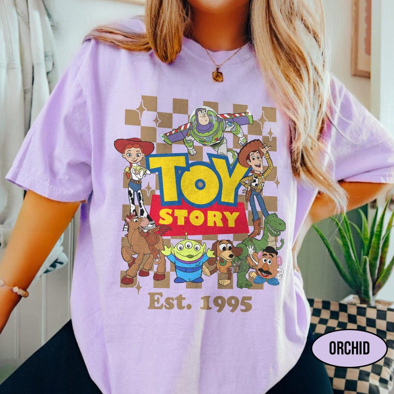 May include: A light purple t-shirt featuring a graphic of characters from the animated film "Toy Story." The design includes the film's title in yellow and red, with the text "Est. 1995" below. The shirt is a comfortable, casual style.