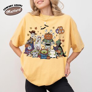 May include: Mustard-coloured t-shirt with a Halloween-themed graphic of animated characters in costumes. The design includes a pumpkin, ghosts, and bats. The words "Comfort Colors" and "Mustard" are printed on the top left.