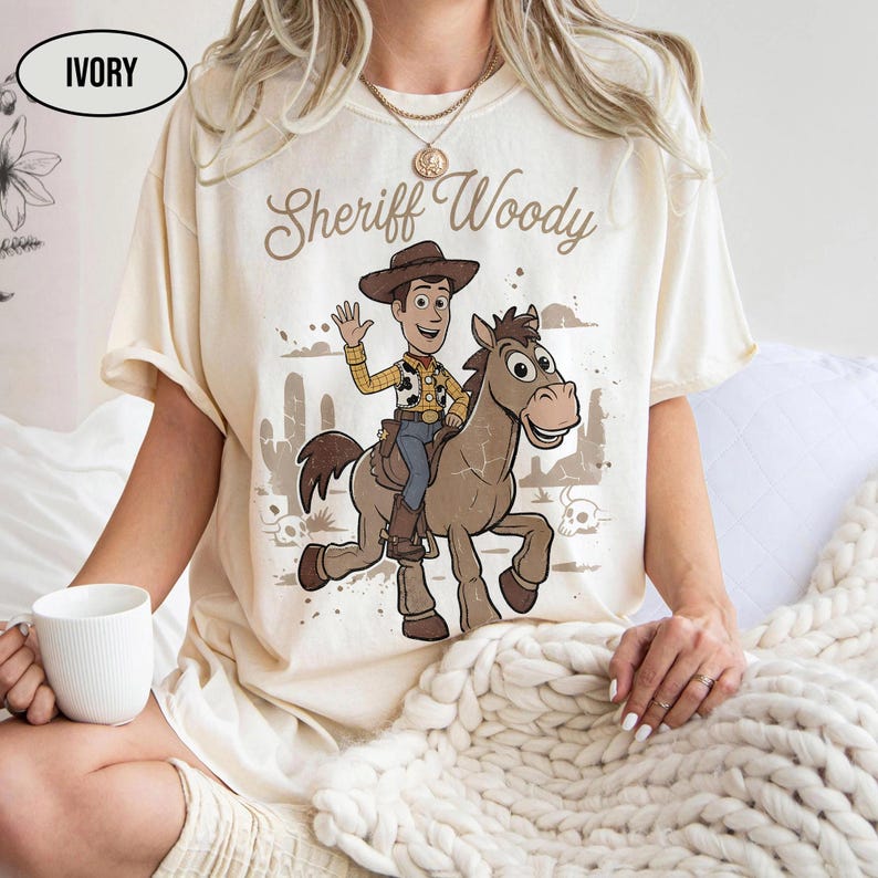 May include: An ivory short-sleeve t-shirt featuring a graphic print of Sheriff Woody riding a horse. The text "Sheriff Woody" is printed above the characters. The design includes a desert background with cacti and skulls in a muted color palette.