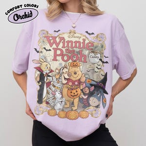May include: A light purple t-shirt featuring a graphic of Winnie the Pooh characters dressed in Halloween costumes. The design includes Pooh, Piglet, Tigger, Eeyore, and Rabbit surrounded by pumpkins, bats, and spiderwebs with the text Winnie the Pooh.