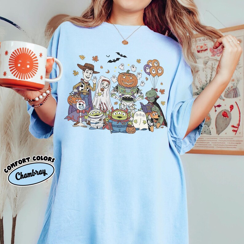 May include: Pale blue t-shirt with a Halloween-themed graphic featuring characters in costumes. The design includes a pumpkin, ghosts, and other characters. The shirt has the words "COMFORT COLORS Chambray" printed on it.