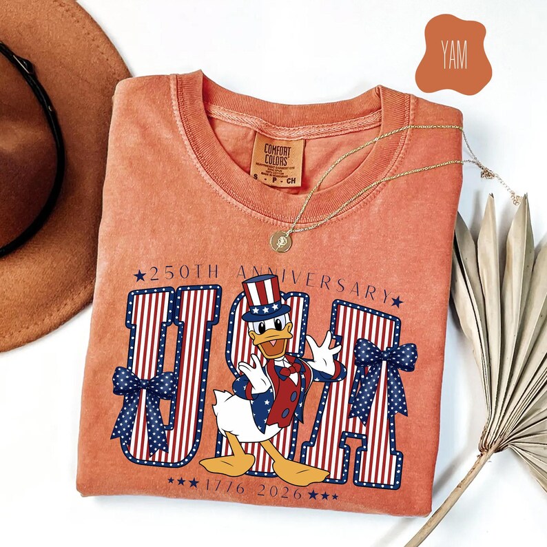 Może przedstawiać: T-shirt z kr&oacute;tkim rękawem w kolorze terakoty z grafiką przedstawiającą kaczkę z kresk&oacute;wki w patriotycznym cylindrze, kamizelce i muszce. Napis głosi 250TH ANNIVERSARY USA 1776 2026, z dużymi czerwono-białymi literami w paski i niebieskimi kokardami.