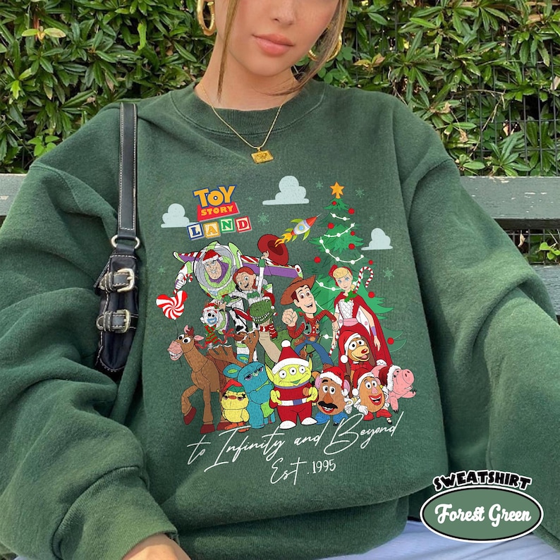 May include: Forest green sweatshirt featuring a colorful graphic of Toy Story characters with a Christmas tree and rocket ship. Text includes "Toy Story Land", "to Infinity and Beyond", and "Est. 1995". The sweatshirt has a crew neck.