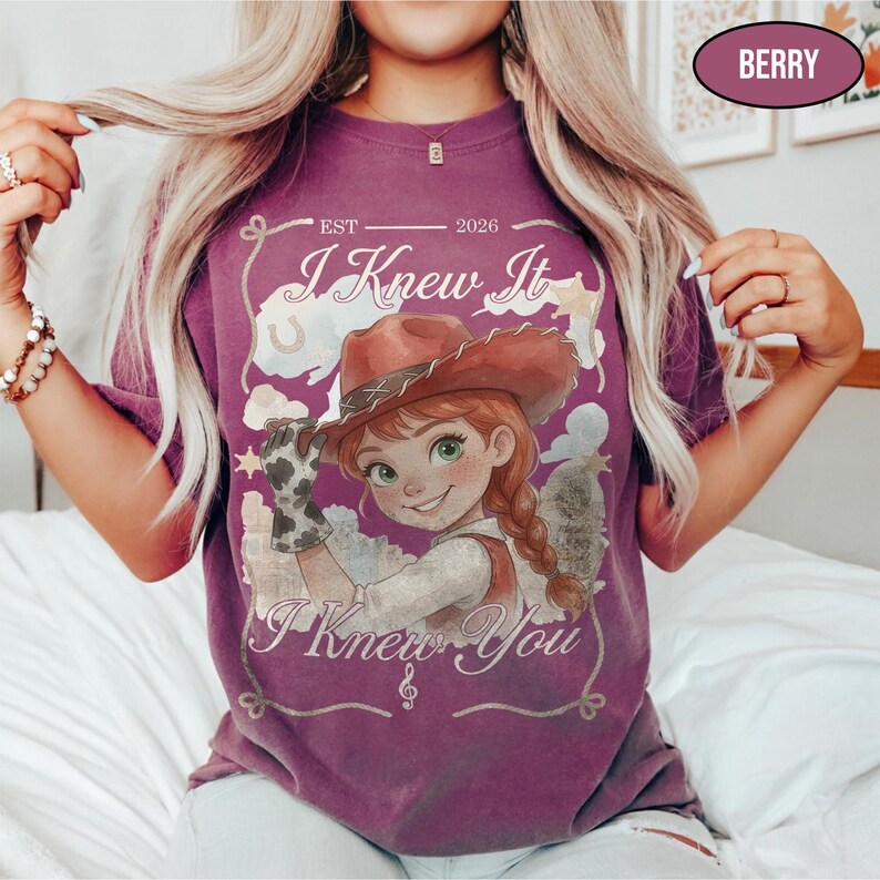 May include: A berry-coloured t-shirt featuring a graphic of a cartoon cowgirl with a braid and a cowboy hat. The text reads "EST 2026", "I Knew It", and "I Knew You". The design includes a lasso border, stars, and a musical note symbol.