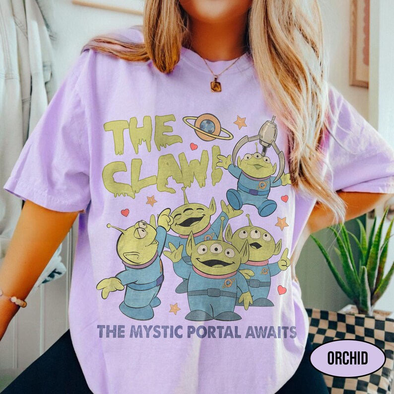 May include: A light purple t-shirt featuring a graphic of green aliens in blue space suits with the text "THE CLAW" and "THE MYSTIC PORTAL AWAITS". The design includes stars, hearts, and a planet. The word "ORCHID" is in a white oval.