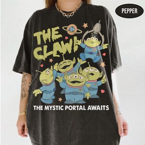 May include: Black t-shirt featuring a graphic of green aliens in blue suits with the text "THE CLAW!" and "THE MYSTIC PORTAL AWAITS". The design includes a planet and stars.
