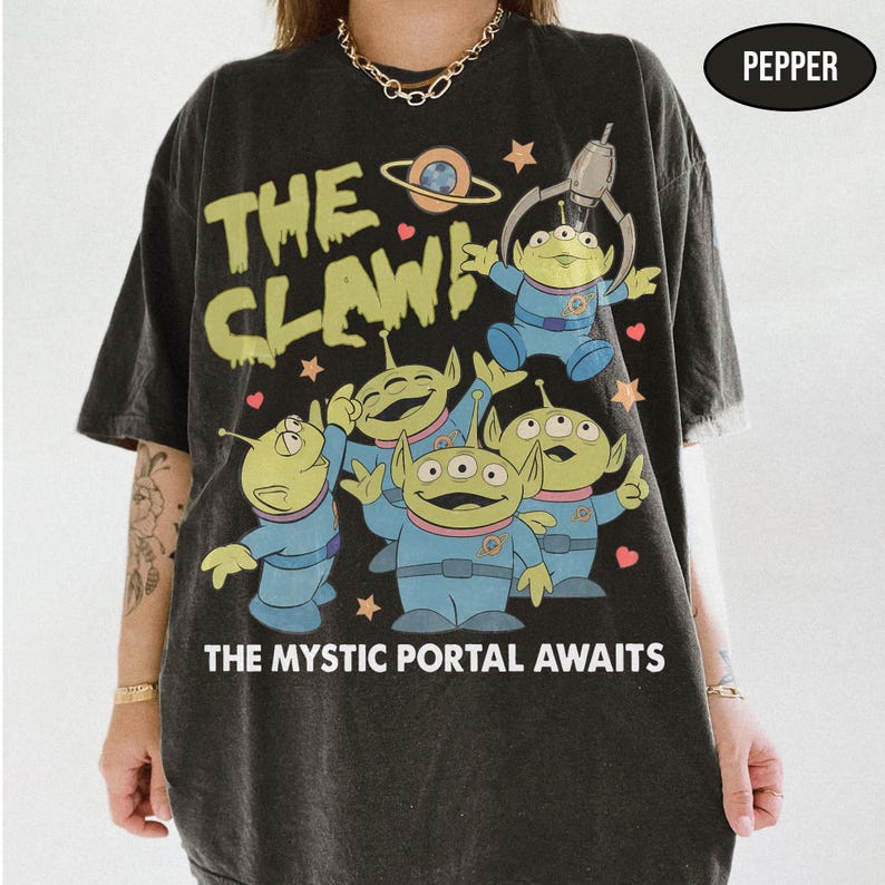 May include: Black t-shirt featuring a graphic of green aliens in blue suits with the text "THE CLAW!" and "THE MYSTIC PORTAL AWAITS". The design includes a planet and stars.
