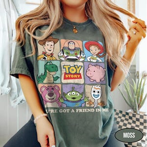 May include: Moss green t-shirt featuring a colorful graphic of Toy Story characters. The design includes Woody, Buzz Lightyear, Jessie, Rex, Hamm, Lotso, the Aliens, and Forky. The text "TOY STORY" and "U'RE GOT A FRIEND IN ME" are also displayed.