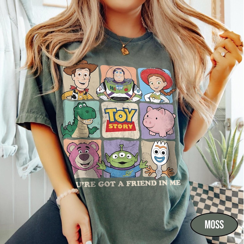 May include: Moss green t-shirt featuring a colorful graphic of Toy Story characters. The design includes Woody, Buzz Lightyear, Jessie, Rex, Hamm, Lotso, the Aliens, and Forky. The text "TOY STORY" and "U'RE GOT A FRIEND IN ME" are also displayed.