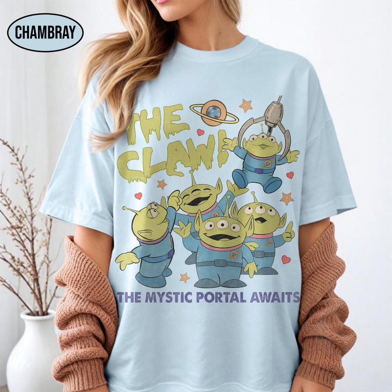 May include: Light blue t-shirt featuring a graphic of green aliens in blue suits with the text "THE CLAW" and "THE MYSTIC PORTAL AWAITS". The aliens are holding a claw machine. The shirt has a crew neck and short sleeves.