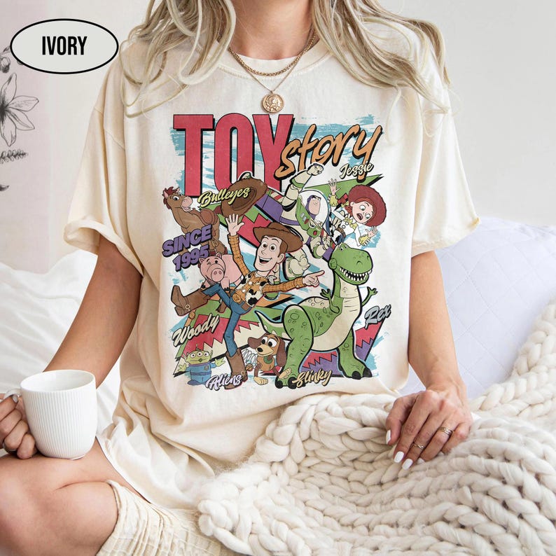 Puede incluir: Camiseta de color marfil con un gr&aacute;fico colorido de Toy Story. El dise&ntilde;o incluye el texto "TOY STORY" y "Since 1995". La camiseta tiene mangas cortas y cuello redondo.