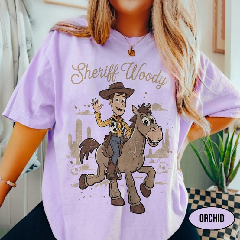 May include: A light purple t-shirt featuring a graphic print of the character Sheriff Woody riding a horse. The text Sheriff Woody is printed in a cursive font above the illustration, which includes desert elements like cacti and skulls.