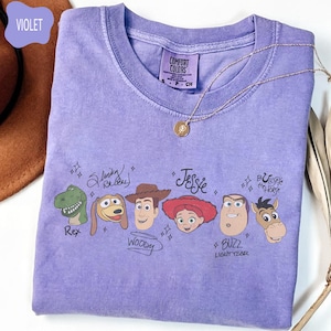 May include: A violet short-sleeve t-shirt featuring a graphic print of Toy Story characters Rex, Slinky Dog, Woody, Jessie, Buzz Lightyear, and Bullseye with their names written in script. The shirt has a Comfort Colors brand label at the neck.