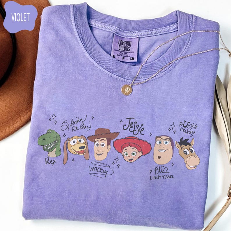 May include: A violet short-sleeve t-shirt featuring a graphic print of Toy Story characters Rex, Slinky Dog, Woody, Jessie, Buzz Lightyear, and Bullseye with their names written in script. The shirt has a Comfort Colors brand label at the neck.