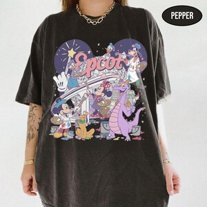 May include: Dark gray t-shirt featuring a colorful graphic of Disney characters painting the word "Epcot". The design includes Mickey Mouse, Minnie Mouse, Donald Duck, Pluto, and Figment. The shirt has a vintage, distressed look.