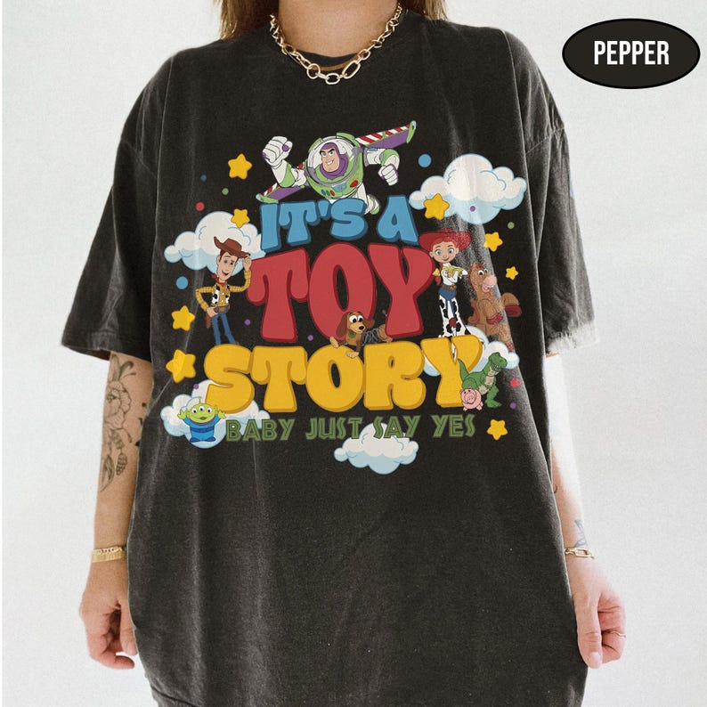 K&ouml;nnte beinhalten: Ein anthrazitfarbenes T-Shirt mit einem Grafikdruck von Toy Story-Figuren wie Buzz Lightyear, Woody und Jessie. Der Text lautet IT'S A TOY STORY BABY JUST SAY YES. Das Design enth&auml;lt bunte Wolken und gelbe Sterne.