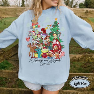 May include: Light blue sweatshirt featuring a colorful graphic of Toy Story characters, a Christmas tree, and the text "Toy Story Land". The phrase "To Infinity and Beyond" and "Est. 1995" are also displayed. The sweatshirt is labeled "Light blue".