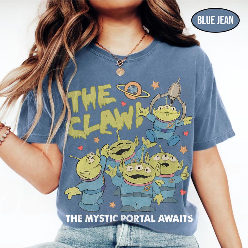 May include: Blue jean-colored t-shirt featuring a graphic of green aliens with three eyes, a claw machine, and the text "THE CLAW!" and "THE MYSTIC PORTAL AWAITS". The design includes stars and hearts.