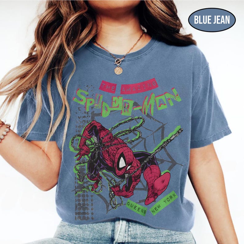 May include: Blue jean colored t-shirt featuring a graphic of Spider-Man in action. The design includes the text "THE AMAZING SPIDER-MAN" in green and red, with "QUEENS NEW YORK" below. The shirt has a vintage, faded look.