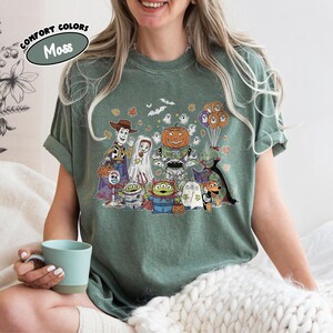 May include: Moss green t-shirt with a Halloween cartoon graphic. The design features a pumpkin, ghosts, and other characters. The words "COMFORT COLORS" and "Moss" are printed on the top left of the shirt. The t-shirt is a comfortable fit.