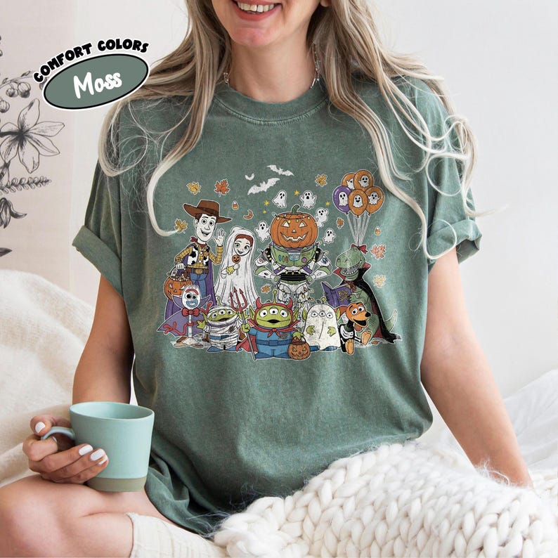 May include: Moss green t-shirt with a Halloween cartoon graphic. The design features a pumpkin, ghosts, and other characters. The words "COMFORT COLORS" and "Moss" are printed on the top left of the shirt. The t-shirt is a comfortable fit.