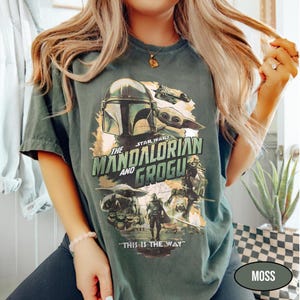 May include: Moss green t-shirt featuring a graphic from "The Mandalorian" with the text "The Mandalorian and Grogu" and "This is the Way." The design includes images of the Mandalorian, Grogu, and other characters from the Star Wars series.