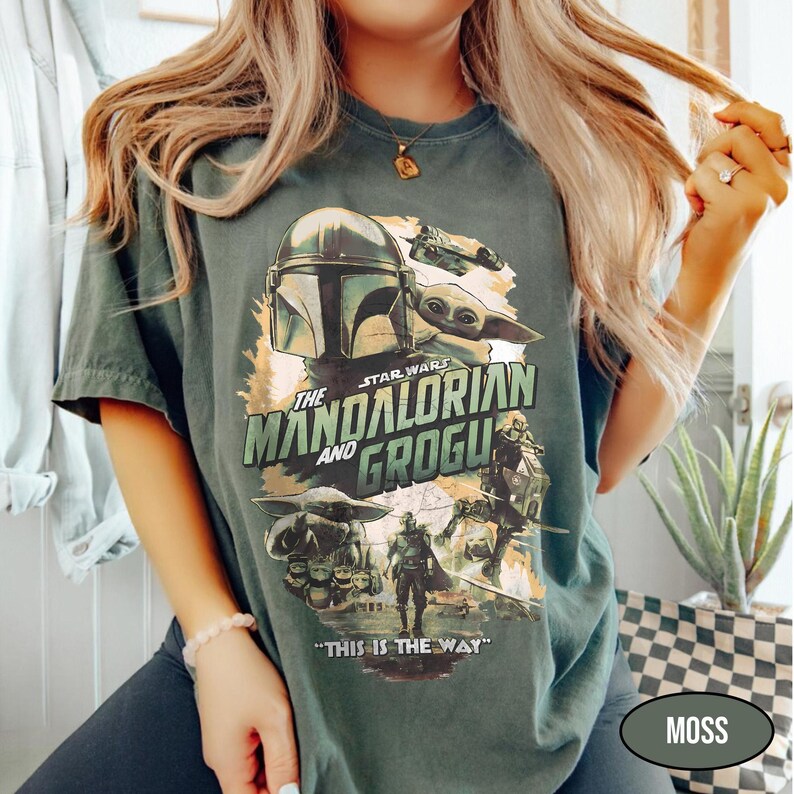 May include: Moss green t-shirt featuring a graphic from "The Mandalorian" with the text "The Mandalorian and Grogu" and "This is the Way." The design includes images of the Mandalorian, Grogu, and other characters from the Star Wars series.